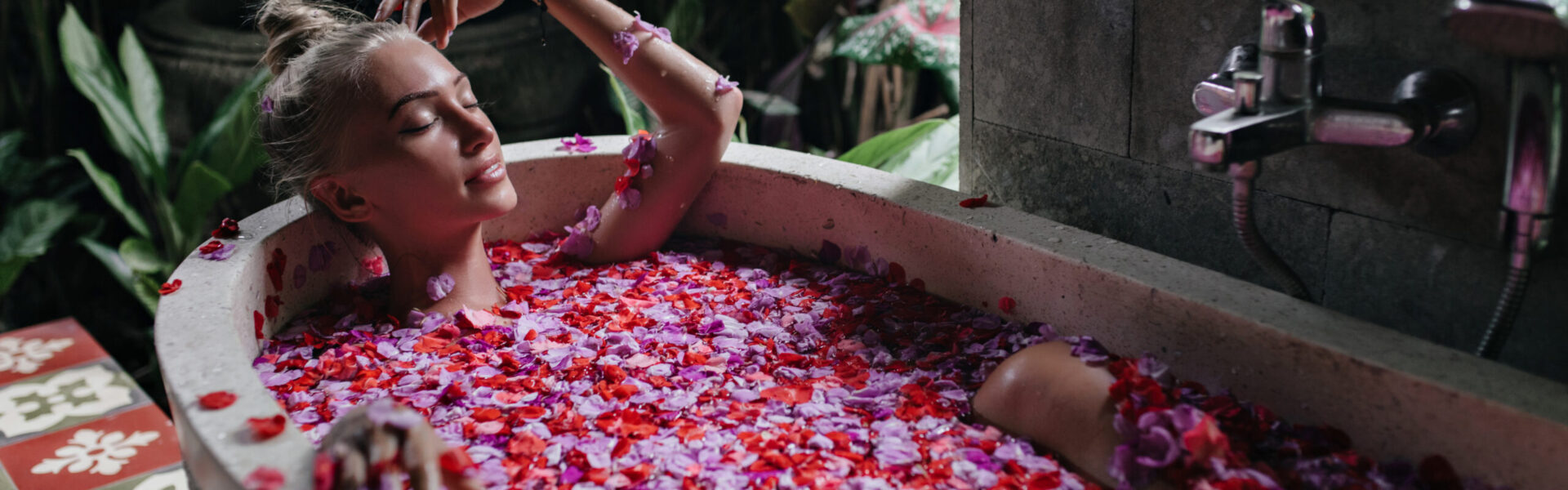 Pleasant woman with tanned skin lying in bath with eyes closed. Indoor shot of cute blonde lady enjoying spa with rose petals.
